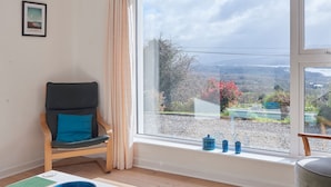 Una habitación con un ventanal grande, una silla, una mesa con un tazón y un cuadro en la pared.
