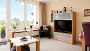 A living room with a large window, a flat-screen TV, a sofa, a table with decorative items, and a chair. - Kurpark- Residenz App. 217 Seesicht, Kurparkallee 47, 27476 Cuxhaven- Döse (Cuxhaven)