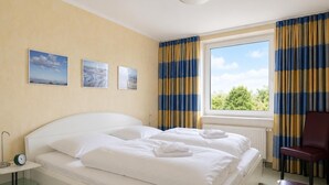 A hotel room with a bed, a chair, a window with curtains, and wall art. - Kurpark- Residenz App. 217 Seesicht, Kurparkallee 47, 27476 Cuxhaven- Döse (Cuxhaven)
