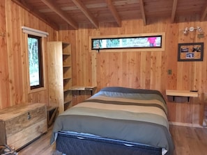 A wooden cabin bedroom with a bed, a dresser, a window, and a shelf with pictures. - Cabaña en el Bosque en la ruta termal (Pichare)