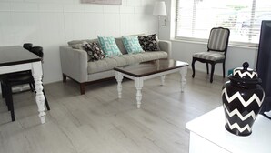 A living room with a sofa, coffee table, and a chair. There is a painting on the wall and a window with blinds. - Hamptons, Port Vincent (Port Vincent)