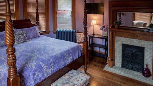 A bedroom with a four-poster bed, a wooden bench, a fireplace, and a radiator.