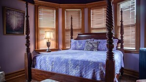 A four-poster bed with a floral-patterned upholstered bench in front, a bedside table with a lamp, and a painting on the wall.