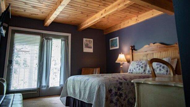 Un dormitorio con techo de madera, una cama con cubrecama floral, una mesita de noche, una lámpara y una ventana con cortinas.