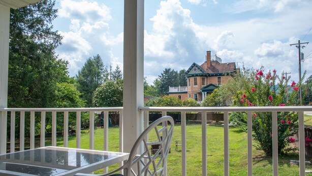 A covered outdoor area with a glass table and metal chair, a grassy lawn, and a brick house in the background.