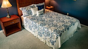 A bedroom with a wooden headboard, a patterned bedspread, two bedside lamps, and a nightstand with a drawer.