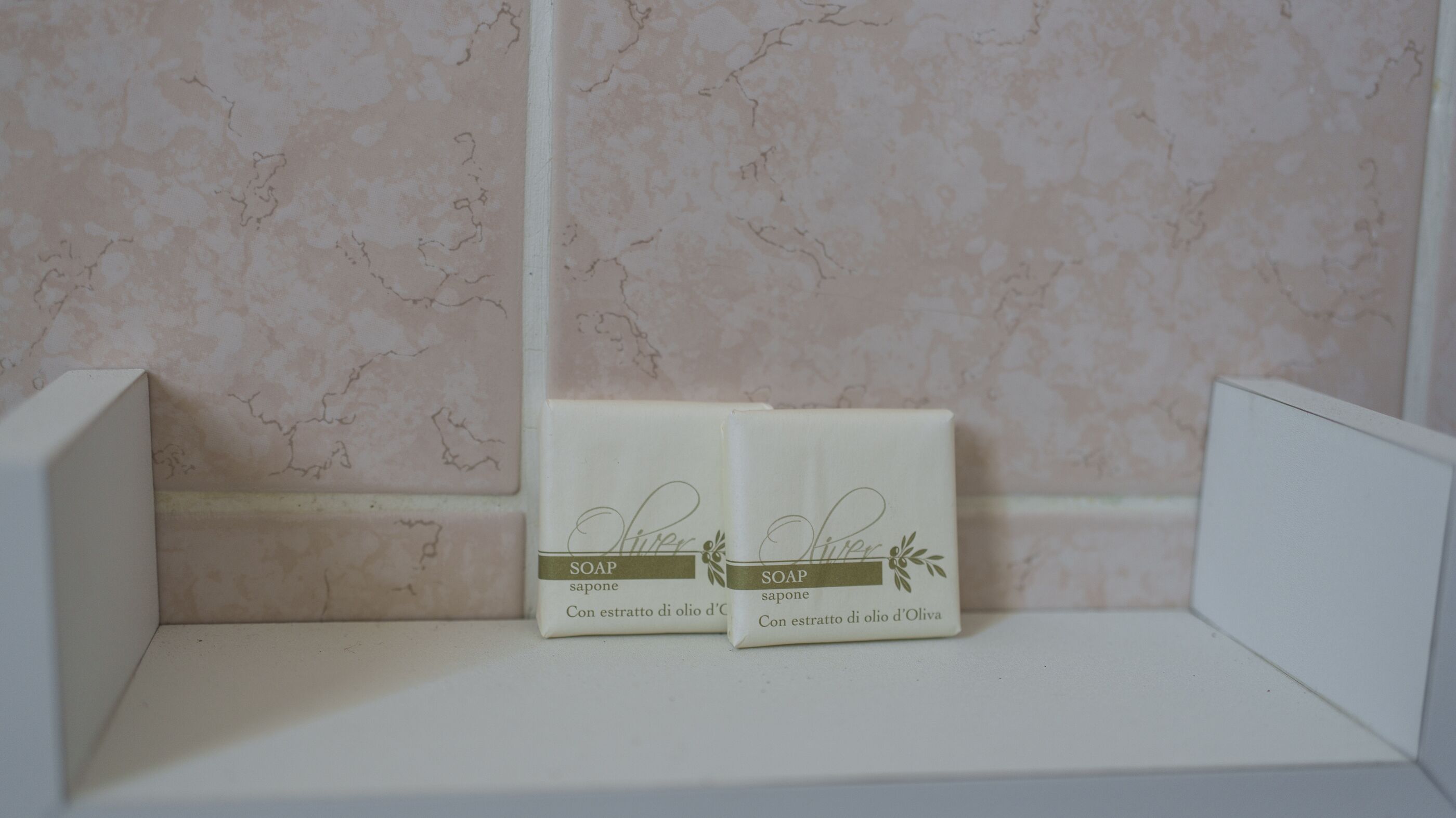 Twee witte zeepstukken met etiketten op een witte plank voor een marmeren muur.