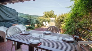 Une terrasse extérieure avec une table dressée pour quatre personnes, entourée de chaises blanches, sous une tonnelle verdoyante.