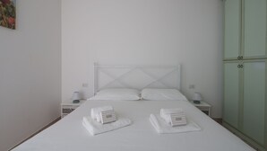 Un lit bien fait, avec du linge de lit blanc et deux serviettes enroulées posées sur le lit. La chambre est équipée de deux tables de chevet avec des lampes et d’une armoire verte.