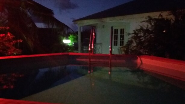 A nighttime view of a swimming pool with a red-lit deck, a white building with a covered entrance, and surrounding vegetation.