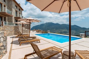 A poolside area with lounge chairs and umbrellas, a stone building, and a mountain view.