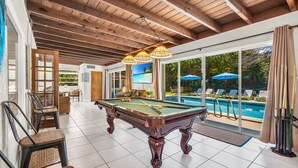 A pool table in a well-lit room with a wooden ceiling and large windows overlooking a pool area. - Birds of Paradise - Historic Spanish Estate (Lake Worth Beach)