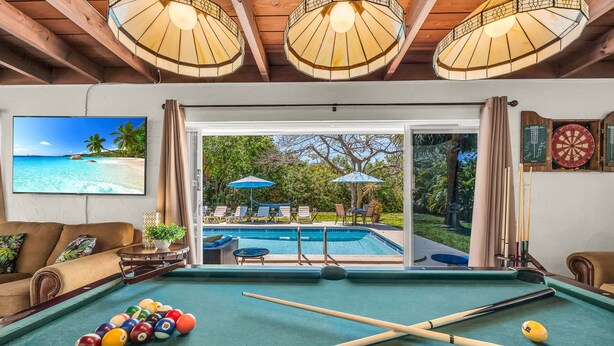 A pool table with pool balls, a television displaying a beach scene, and a view of a pool and outdoor seating area. - Birds of Paradise - Historic Spanish Estate (Lake Worth Beach)