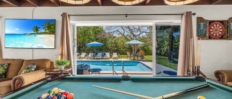 Une table de billard avec des billes, un téléviseur diffusant une scène de plage et une vue sur une piscine et un espace de détente extérieur.