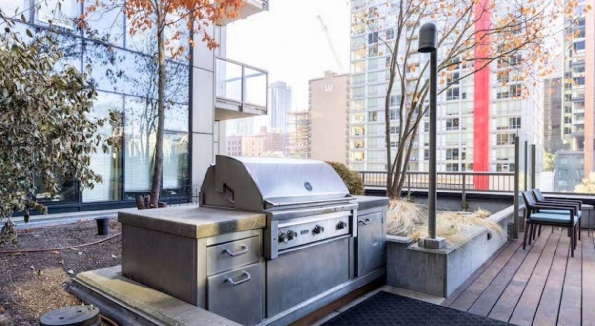 An outdoor grill area with a stainless steel grill and seating, surrounded by a wooden deck and a view of tall buildings.