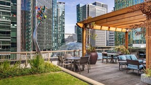 An outdoor terrace with wooden furniture and a pergola, overlooking a cityscape with modern buildings. - Luxury Living: 2BR Condo at Via 6 (Seattle)