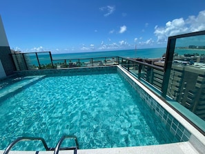 A rooftop infinity pool with clear blue water, surrounded by a glass railing, offering a view of the ocean and cityscape.