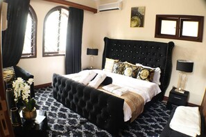 Un dormitorio con una cama grande, dos mesitas de noche, una mesita auxiliar, un ventanal con cortinas y un equipo de aire acondicionado instalado en la pared.