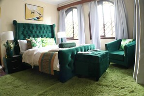 Un dormitorio con una cama tapizada de verde, un sillón verde y un reposapiés verde. Se ve una ventana con cortinas blancas y un cuadro en la pared.