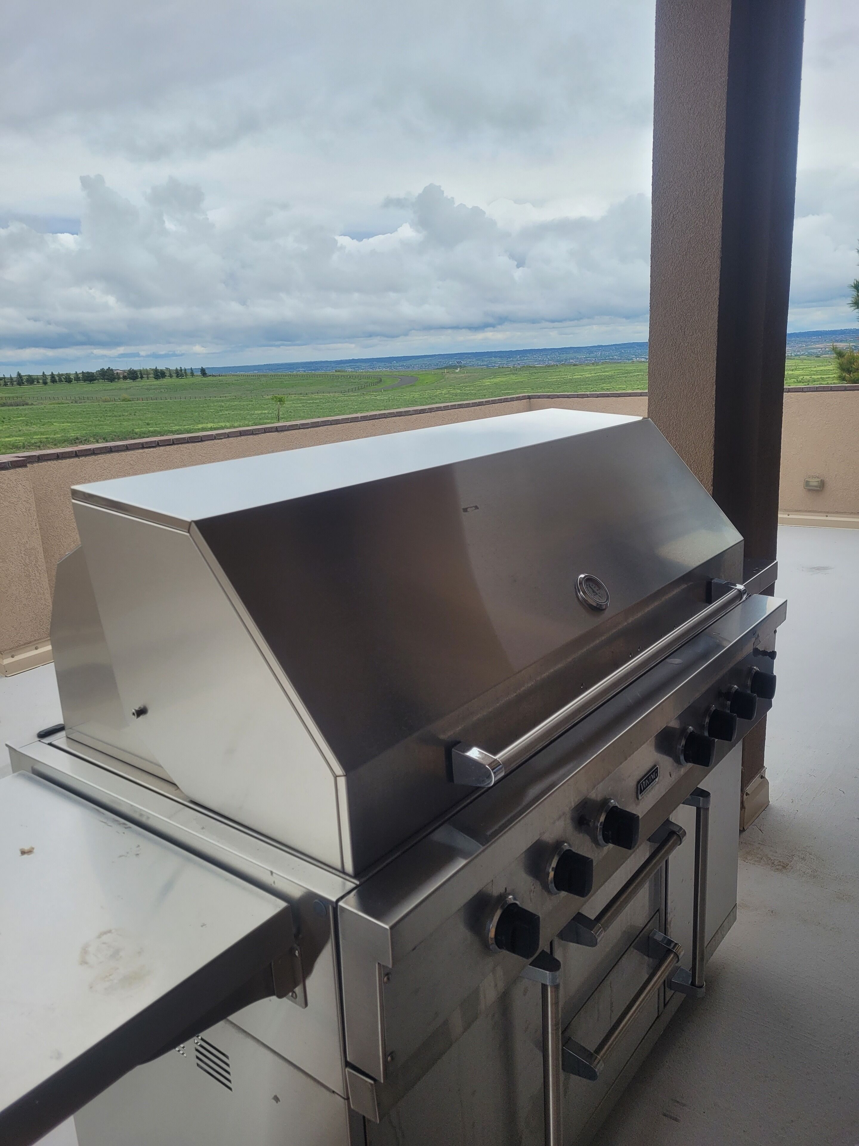 A stainless steel barbecue grill with a flat top and multiple burners, positioned on an outdoor patio with a view of a green field and cloudy sky.