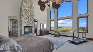 A spacious bedroom with a large bed, a stone fireplace, a treadmill, and a view of the outdoors. - Luxury Mansion Retreat, Sleeps 16+ ,Panoramic VIEWS 36 Acre Land .in the city . (Castle Rock)