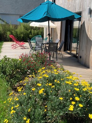 Un patio con tavolo e sedie, circondato da un giardino con fiori gialli e un ombrellone blu.