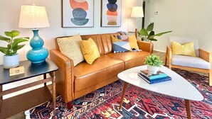 A living room with a brown leather sofa, a white coffee table, and a patterned rug. There are two lamps on side tables, a blue vase, and a small wooden box. - The Leia 2BR Bright Retreat by Velvet Ditch Villas (Oxford)