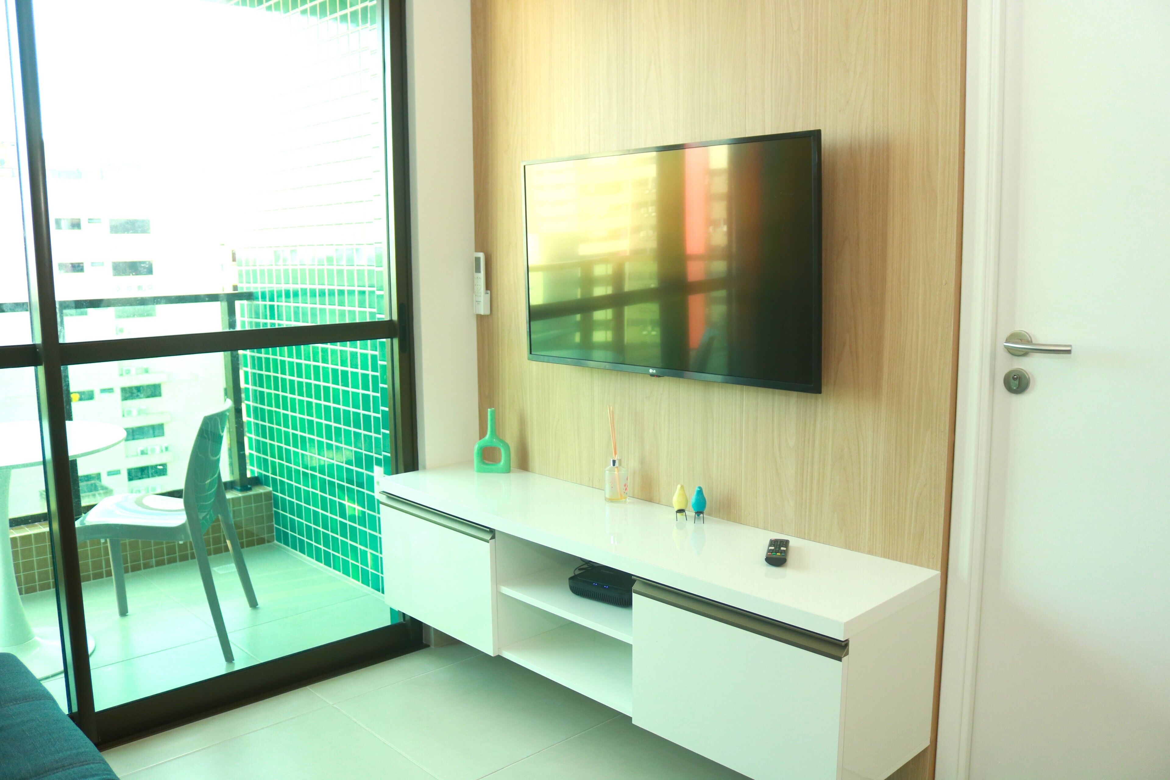 A modern room with a large TV mounted on a wooden wall, a white desk with a chair, and a view through a glass door to an outdoor area with a green fence and a table.