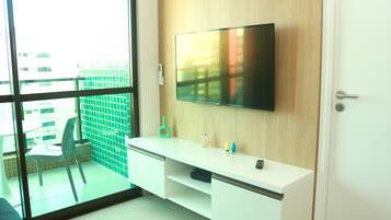 A modern room with a large TV mounted on a wooden wall, a white desk with a chair, and a view through a glass door to an outdoor area with a green fence and a table.