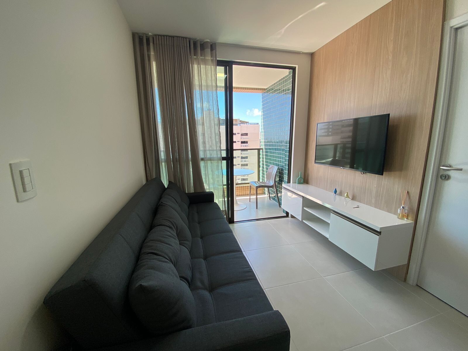A modern living room with a black sofa, a white TV stand, and a sliding glass door leading to a balcony.