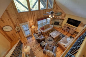 A cozy cabin interior with a fireplace, leather sofas, a television, and a large window overlooking the outdoors. - Big Sky Overlook - Hottub- Firepit - Pool Table (Blairsville)