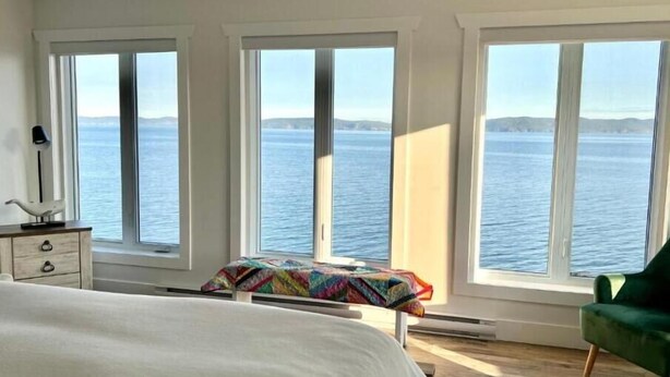 Une chambre à coucher avec un grand lit, un fauteuil vert et une vue sur la mer, visible à travers trois fenêtres.