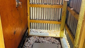 Una puerta de madera con un picaporte metálico, una valla de madera con listones horizontales y un camino de grava que conduce a una puerta.