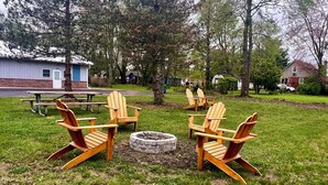 Un espacio al aire libre con césped, sillas Adirondack de madera dispuestas alrededor de un hogar de piedra.