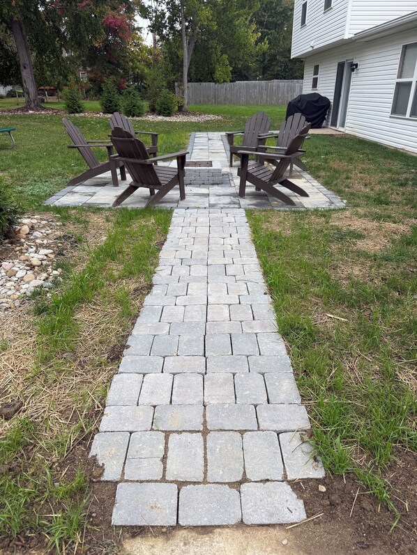 Un patio empedrado con sillas Adirondack y una mesa, rodeado de un jardín césped y una valla de madera.