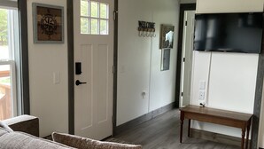 A modern living room with a dark wood floor, a round coffee table, and a sofa. There is a white door with a window, a wall-mounted TV, and a 'WELCOME' sign. - Cozy little tiny home close to Whitefish, Columbia Falls, and Airport (Columbia Falls)