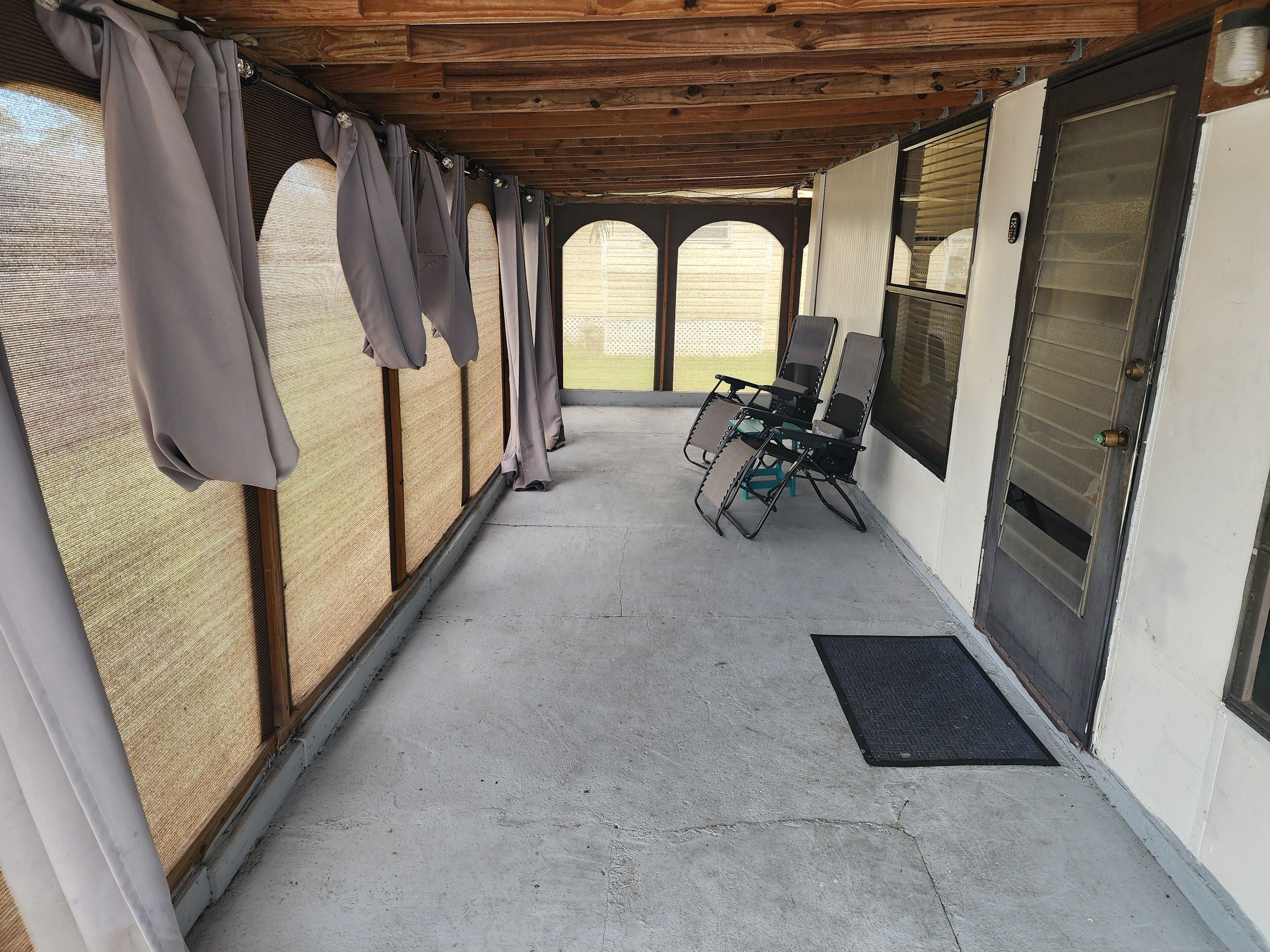 A covered outdoor area with a concrete floor, a door with a metal handle, and a window with a metal grill.