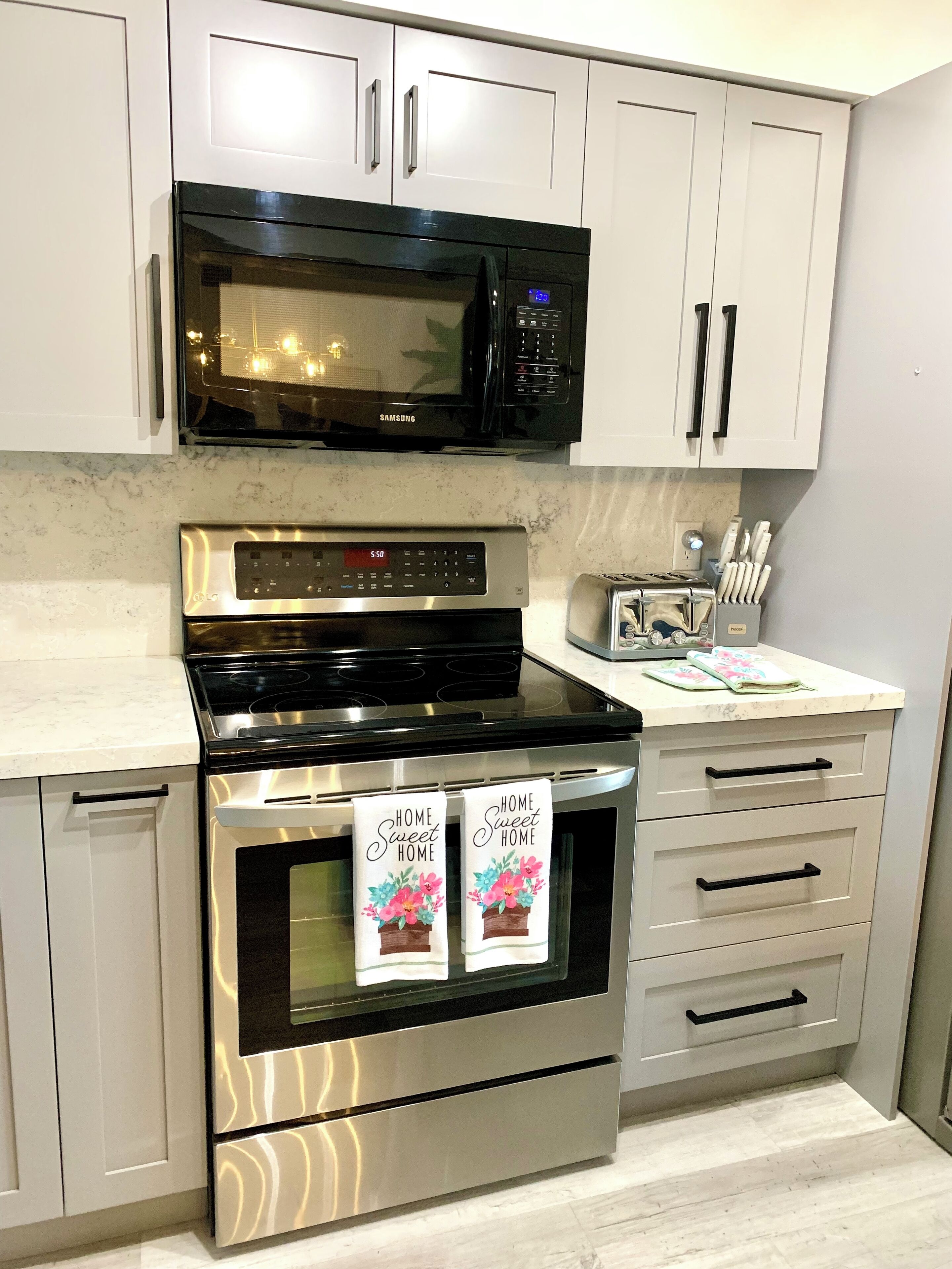 A modern kitchen with stainless steel appliances, including a microwave, oven, and toaster. There are two towels hanging on the oven handles with the text "HOME Sweet HOME".