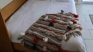 A bedroom with a wooden bed frame, white bedding, a decorative throw blanket, and a small shelf with items on the wall. - Adorable Gun Lake Cottage (Shelbyville)