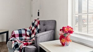 Una habitación con un sillón tapizado gris, una mesita con un florero y una ventana con persianas.