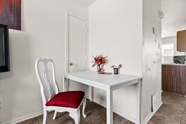 Un pequeño comedor con una mesa blanca, una silla con cojín rojo y un florero con flores.