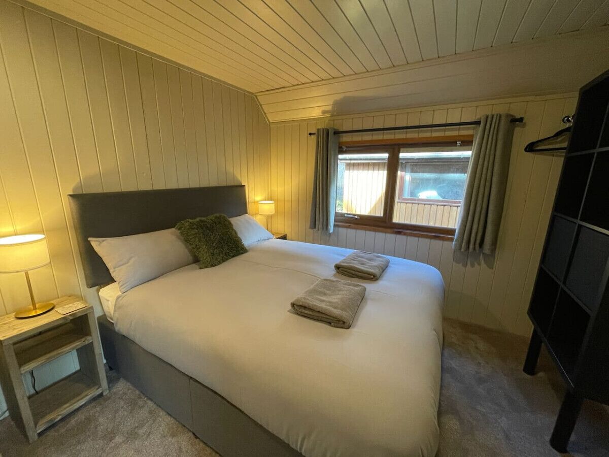 Een keurig opgemaakt bed met twee kussens en twee handdoeken, een nachtkastje met een lamp, en een raam met gordijnen.