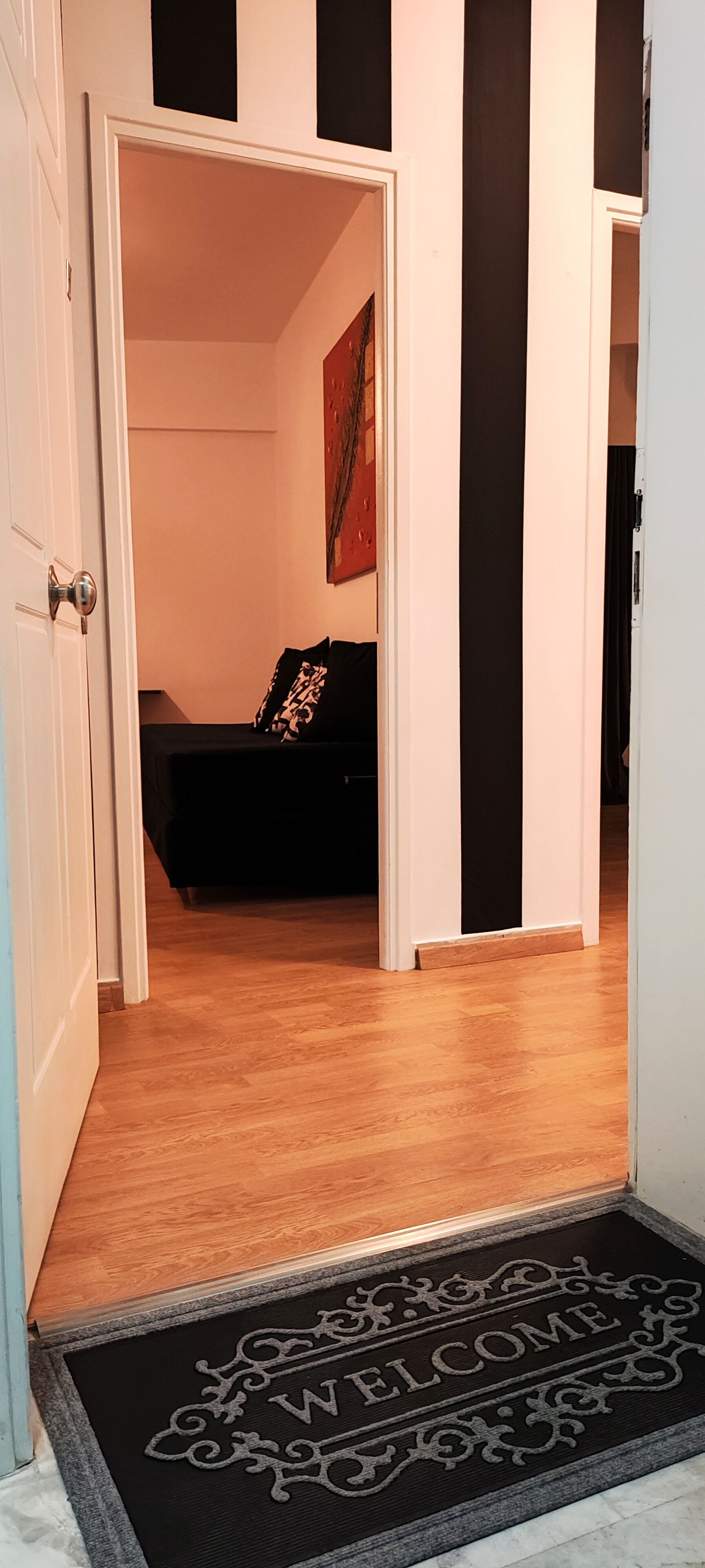 A hallway with a doormat reading "WELCOME" leading to a room with a bed and a painting on the wall.