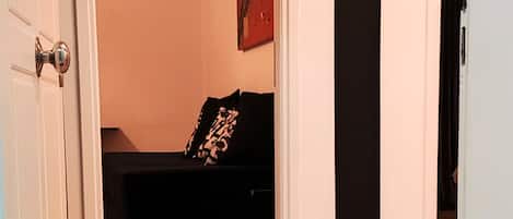 A hallway with a doormat reading "WELCOME" leading to a room with a bed and a painting on the wall.