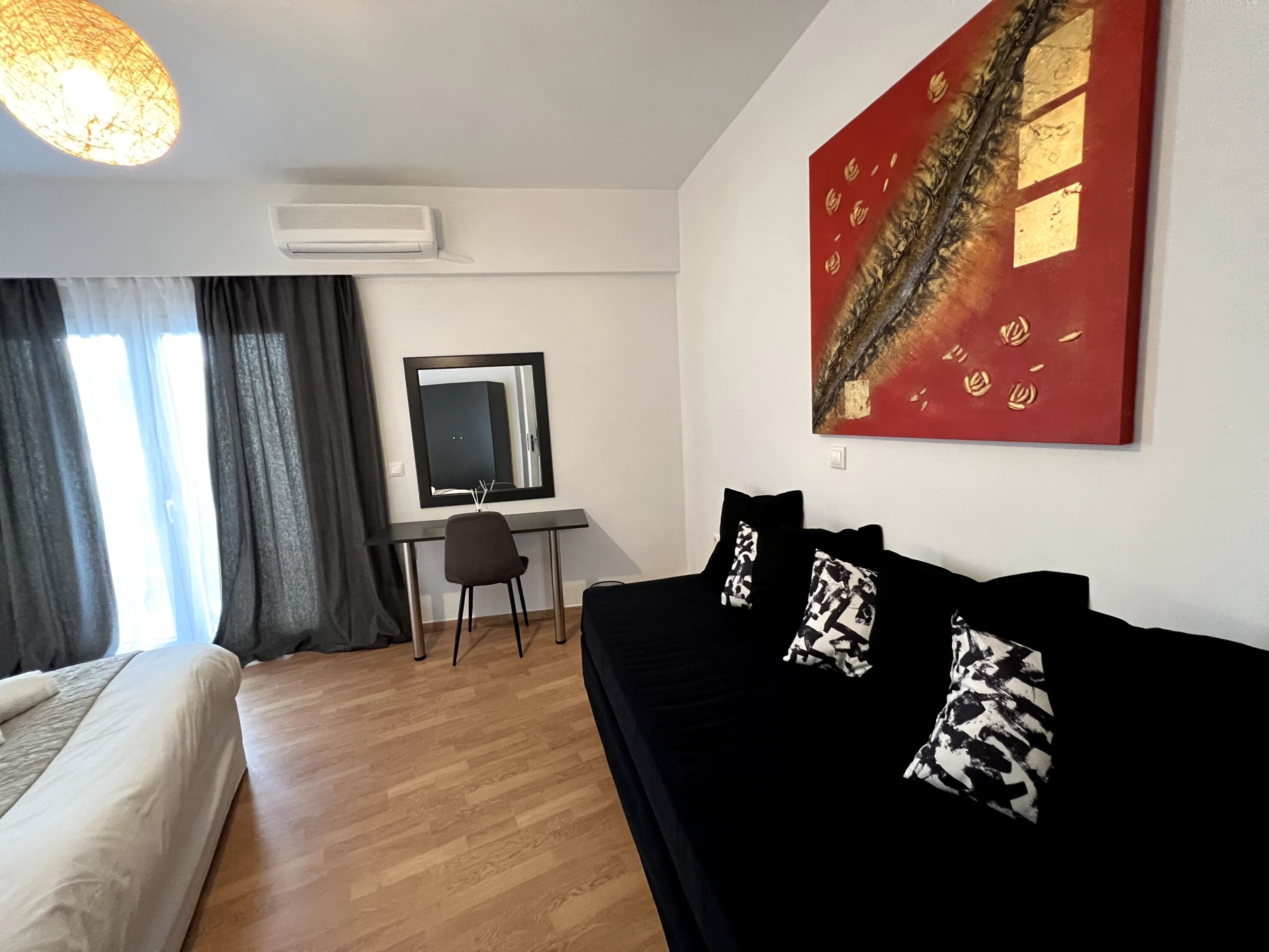 Una camera d'albergo moderna con un letto, un divano nero con cuscini decorati, una scrivania con una sedia, un quadro astratto di grandi dimensioni e una finestra con tende.