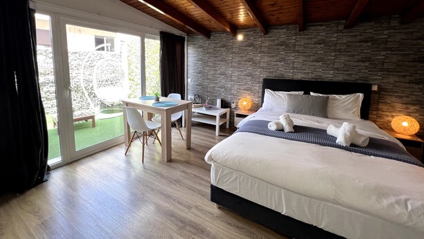 A bedroom with a large bed, a dining table, and a view of a garden through glass doors. - Allure Athens Urban Suites (Athens)