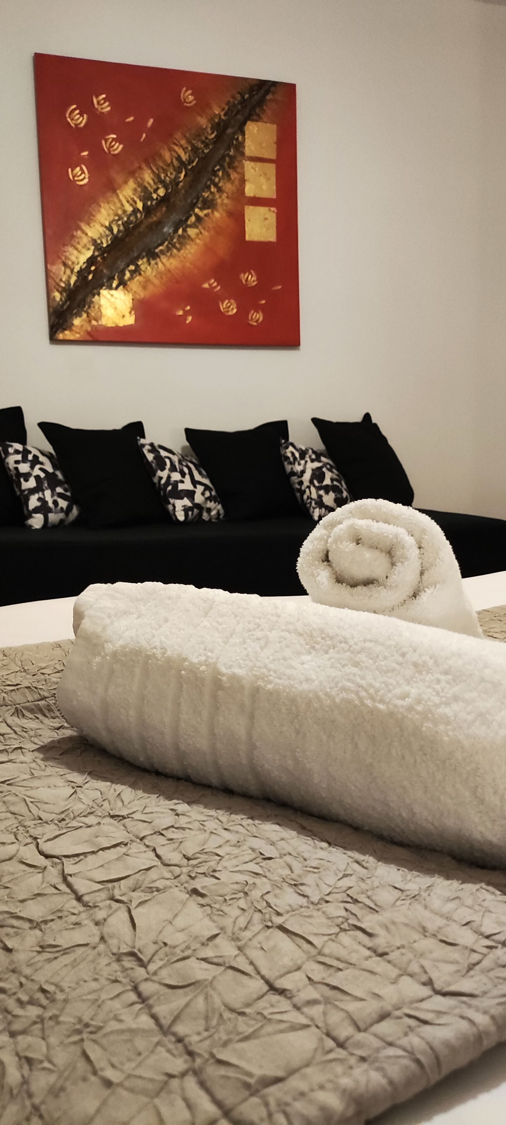 A neatly made bed with a rolled white towel, a patterned blanket, and a red abstract painting on the wall.