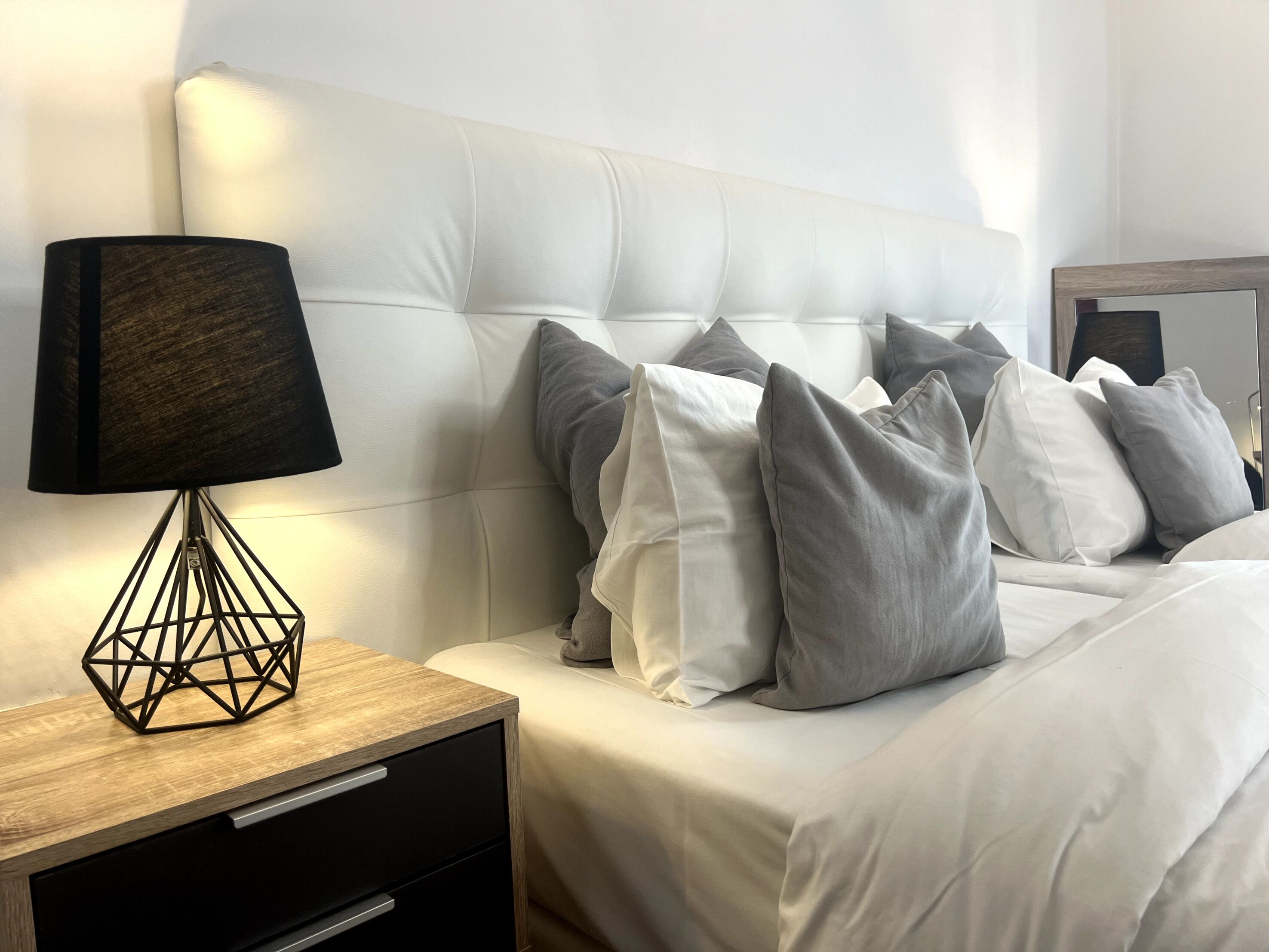 A neatly made bed with white and gray pillows, a wooden nightstand with a geometric lamp, and a mirror on the wall.