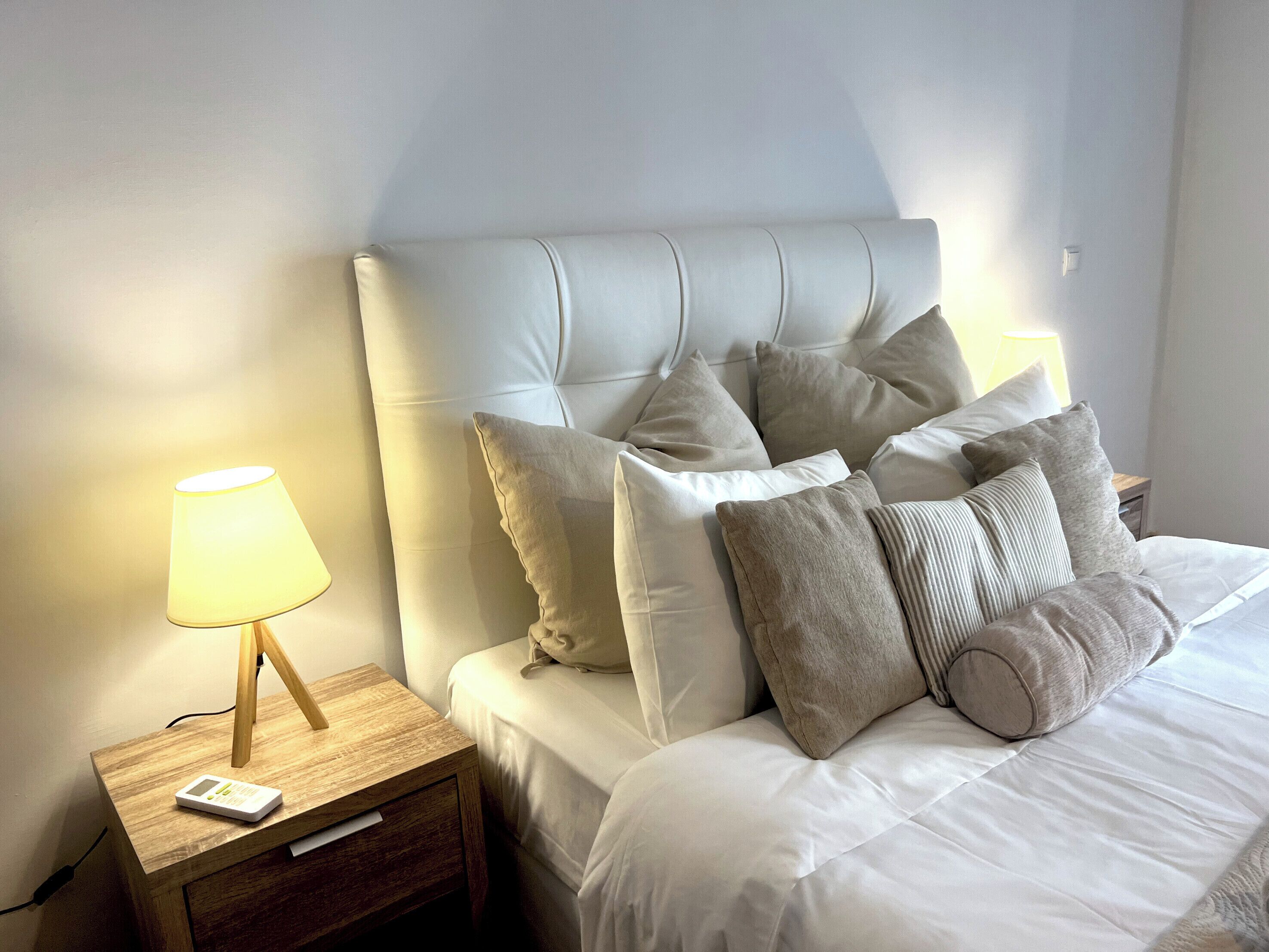 Ein ordentlich bezogenes Bett mit Kopfteil, mehreren Kissen, einer Nachttischlampe und einem Nachttisch aus Holz mit einem digitalen Gerät.