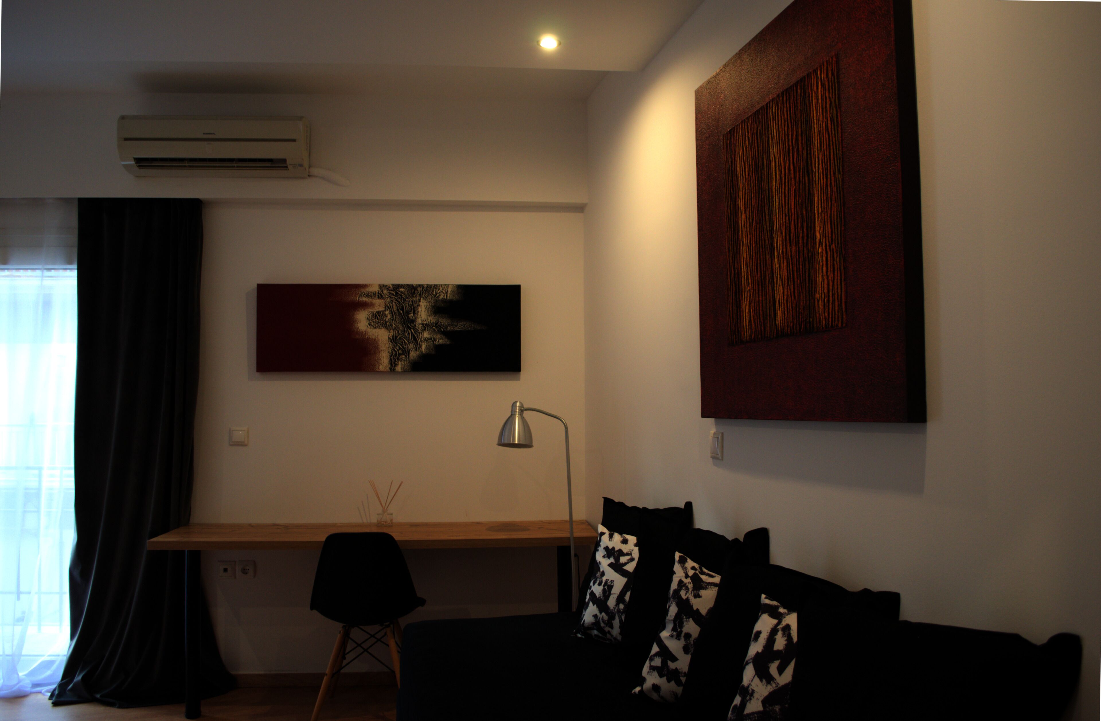 A modern living room with a wooden desk, a black couch with patterned cushions, and two abstract paintings on the walls.